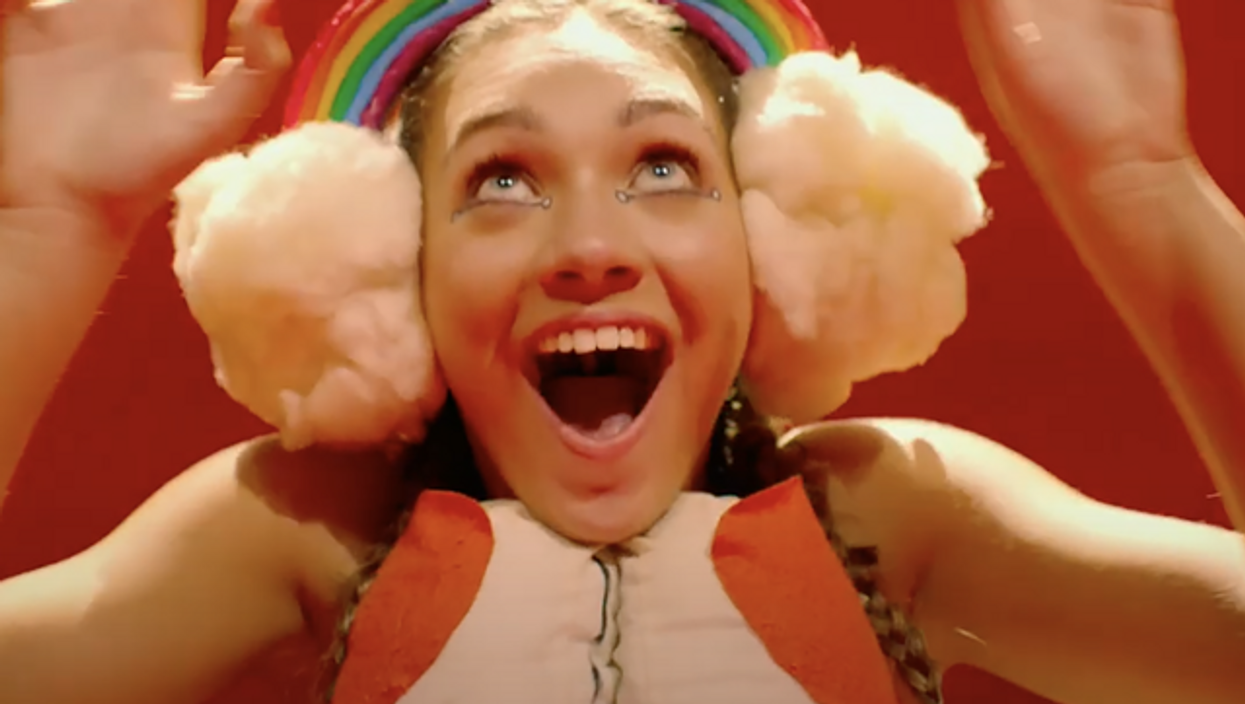 Maddie Ziegler in the film Music. Her mouth and eyes are opened wide, and she is smiling. On her head is a rainbow, with two clouds either side which look like earmuffs.