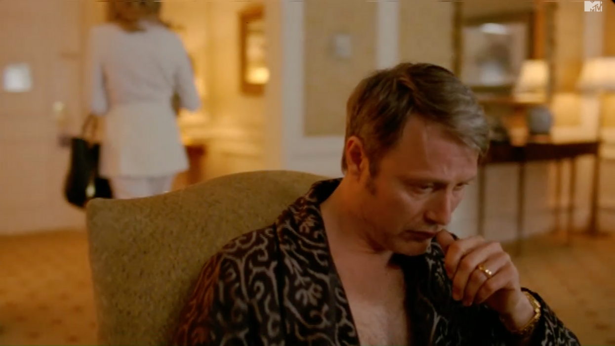 Mads Mikkelsen in a black dressing gown places a thumb to his mouth, he’s in an armchair, thinking deeply. Behind him, a women in white heads to the door.