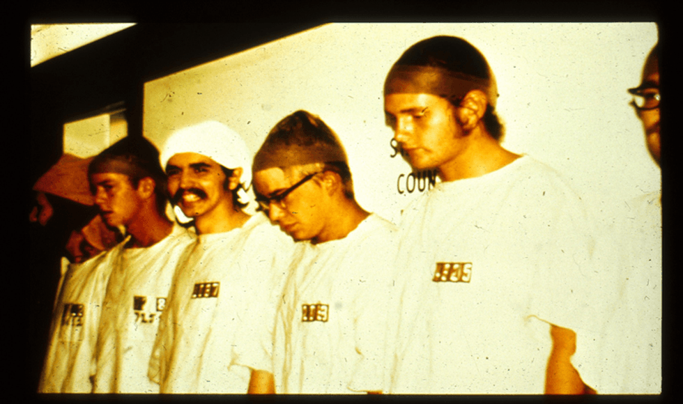 Male participants in the Stanford Prison Experiment stand against a white wall wearing white prisoner T-shirts.