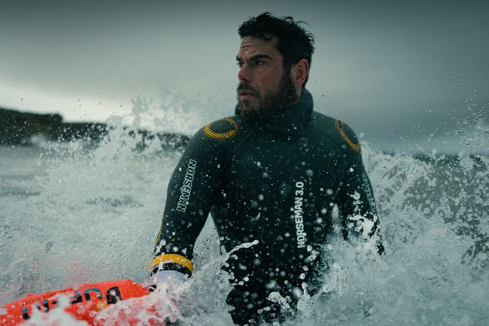 Man in a wetsuit with the waves crashing behind him as he looks away from the camera