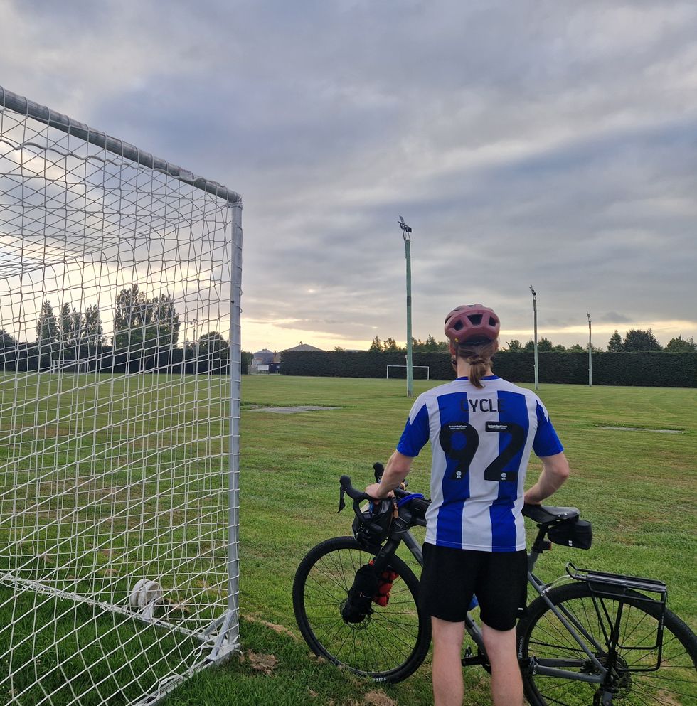 Man wearing a custom football shirt with the words 'Cycle 92' as he wears a cycling helmet and is stood with his bike