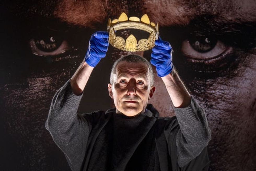 Man wearing gloves holding a crown, with a pair of eyes on a large poster behind him