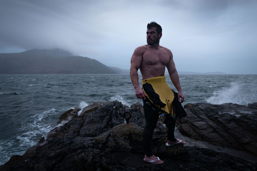 Man wearing wetsuit to his waist with his bare torso exposed as he stands on rocks with the sea behind him