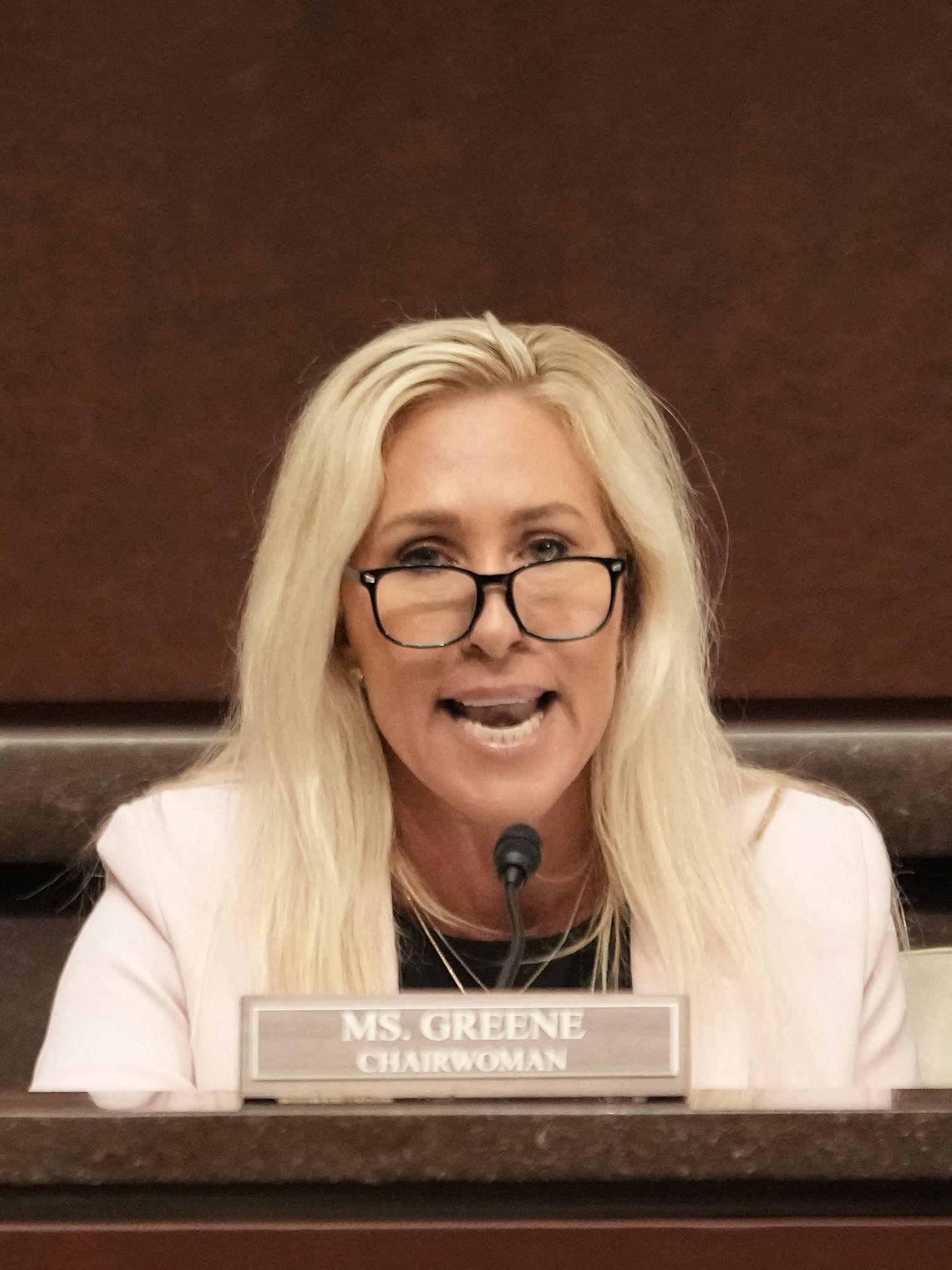Marjorie Taylor Greene, a white woman with glasses and long blonde hair, speaks behind a desk with a sign reading 'Ms. Greene. Chairwoman'.