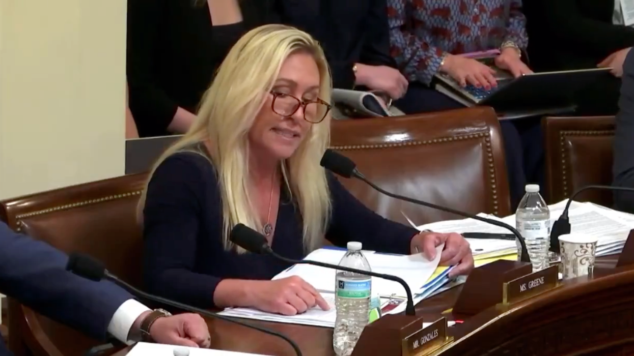 Marjorie Taylor Greene, a white woman with long blonde hair and glasses, reading from notes in front of her on a committee bench.