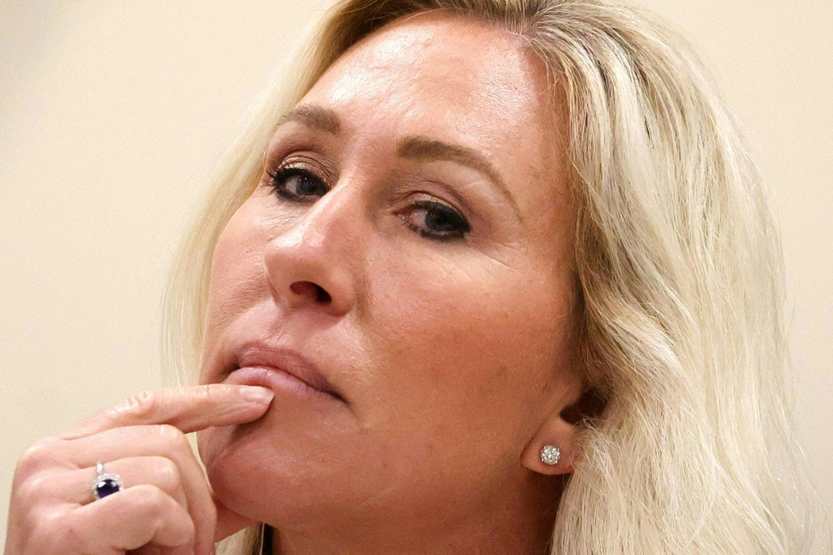 Marjorie Taylor Greene, a white woman with long blonde hair, places her index finger on her chin, in thought, with her head tilted back slightly.