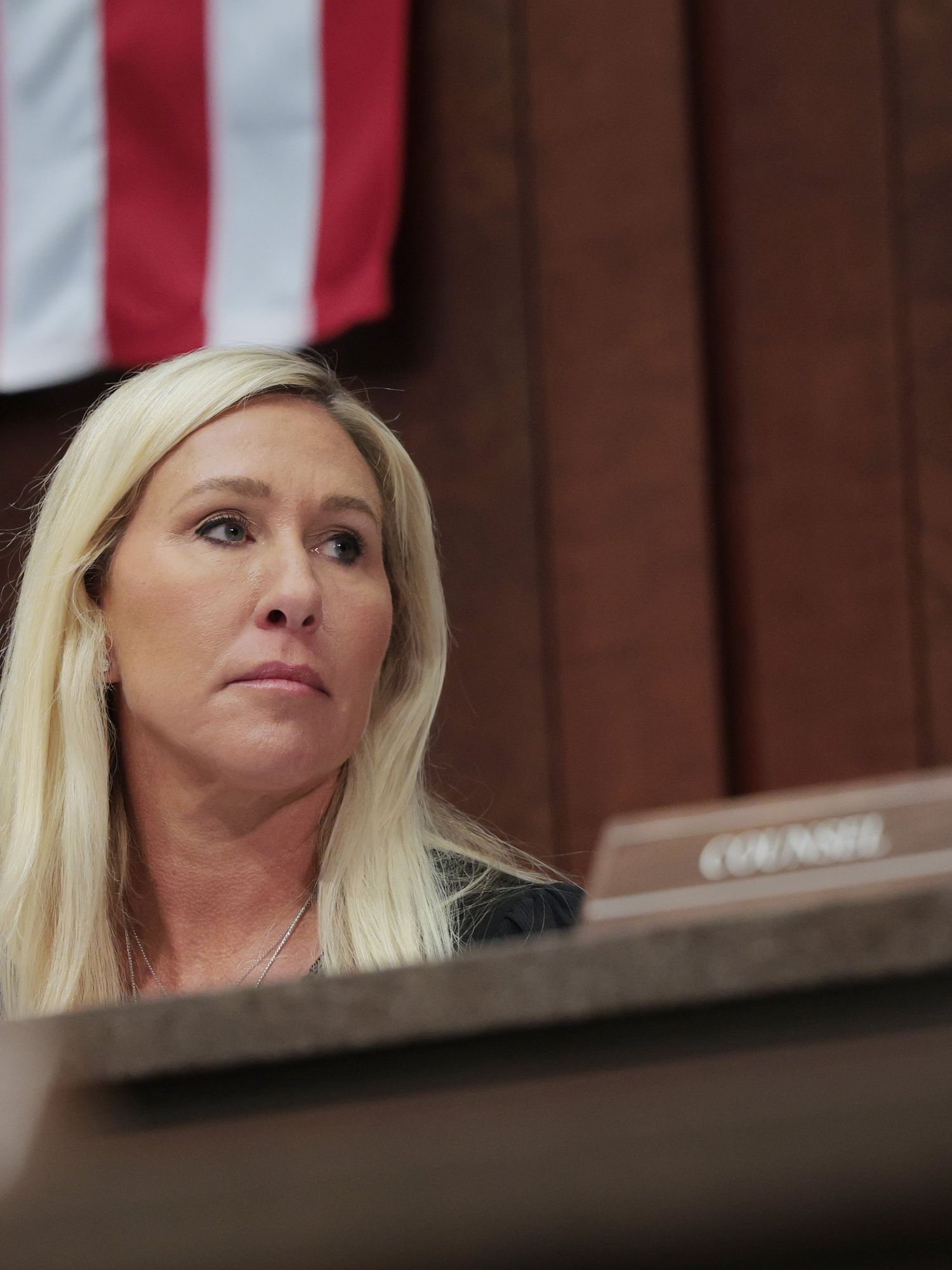 Marjorie Taylor Greene, a white woman with long blonde hair, sitting on a committee and looking to her left with a neutral expression on her face.