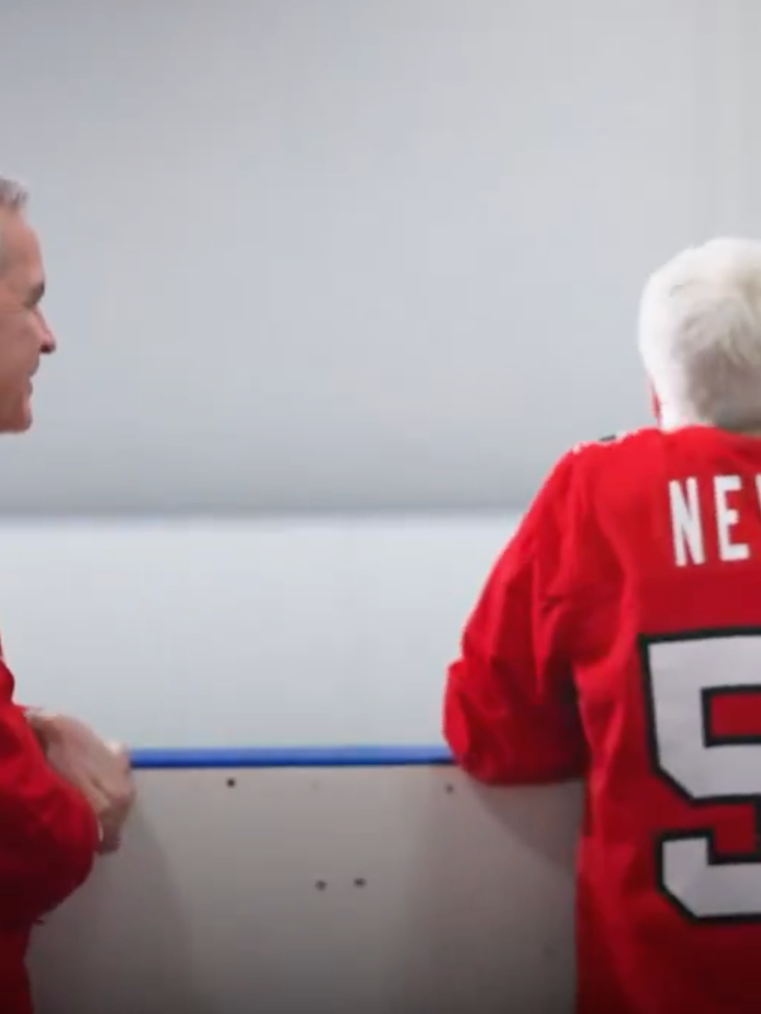 Mark Carney, left, and Mike Myers, right, watch an ice hockey game from the sidelines. Myers has his back to the camera, with his red hockey shirt reading: 'Never 51'.