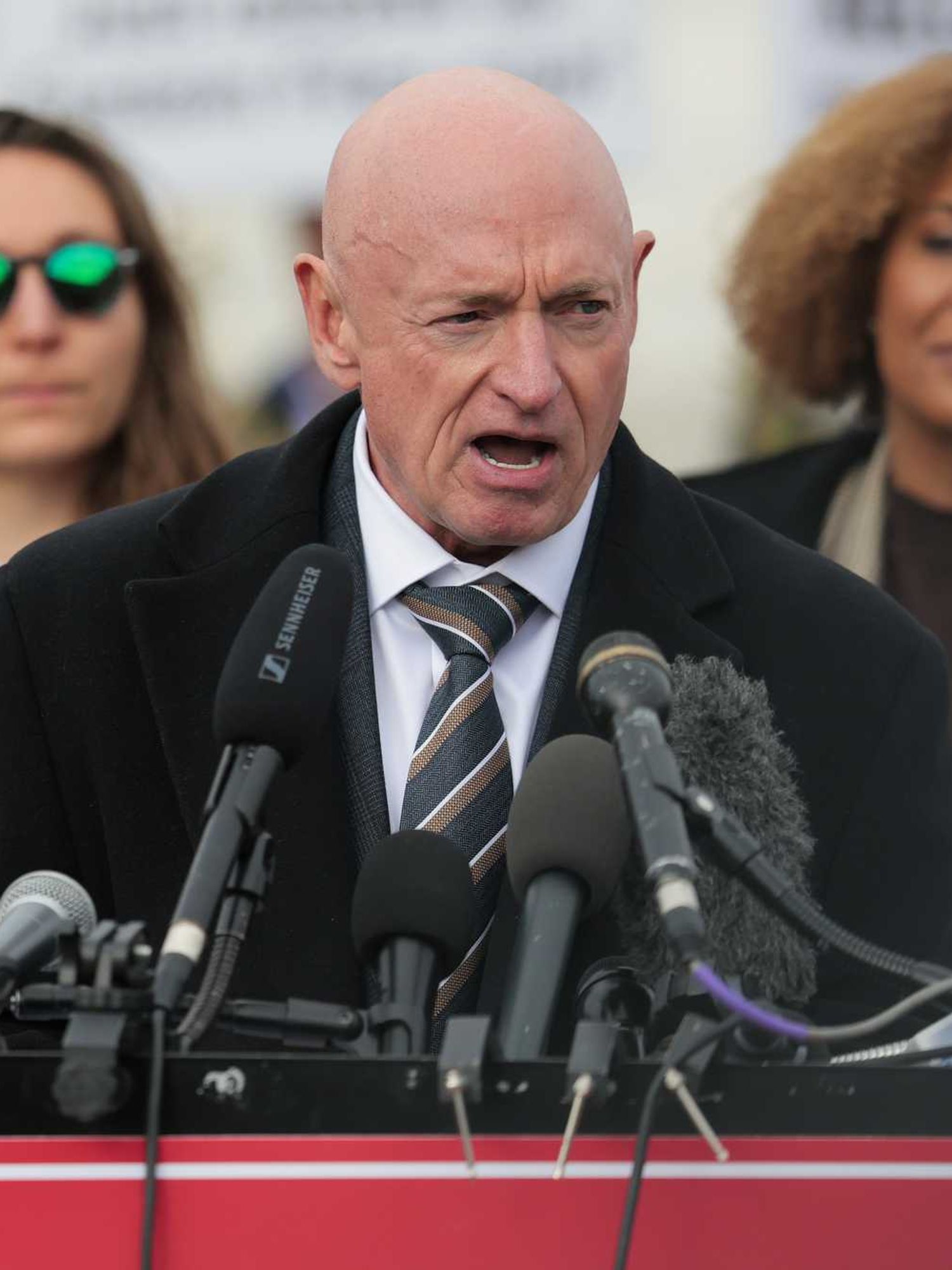 Mark Kelly, a bald white man, speaking into several microphones on a lectern.
