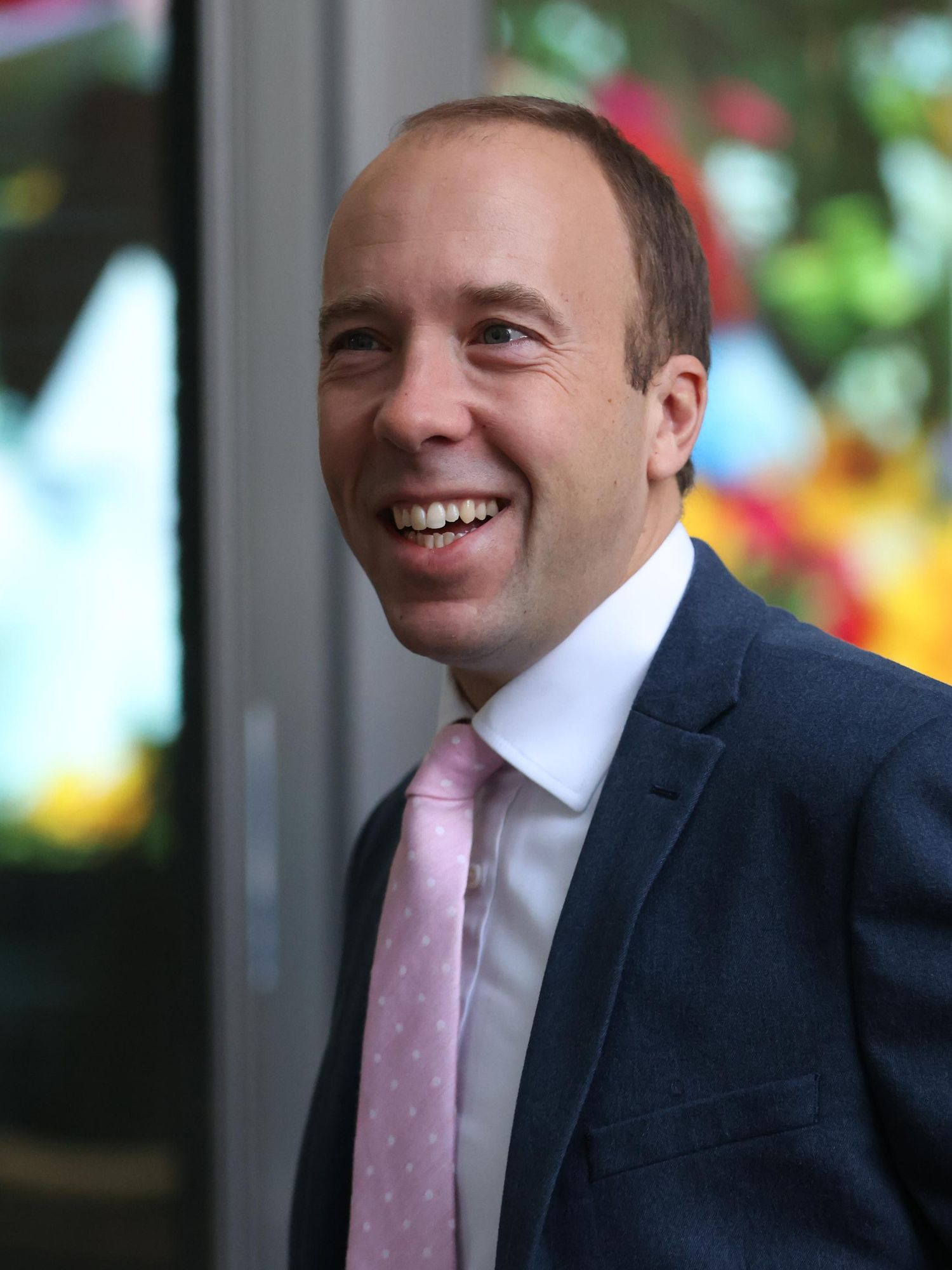 Matt Hancock, a white man with a large forehead, short brown hair and a navy suit, smiles widely at someone off-camera as he walks past a building.