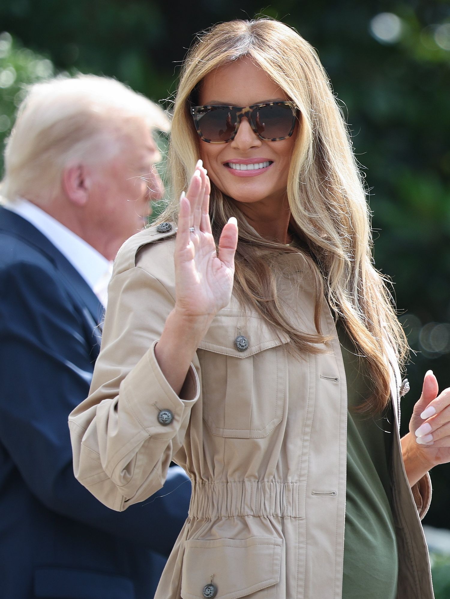 Melania Trump, with sunglasses and wearing a cream jacket, smiles and waves at reporters, with Donald Trump in the background.