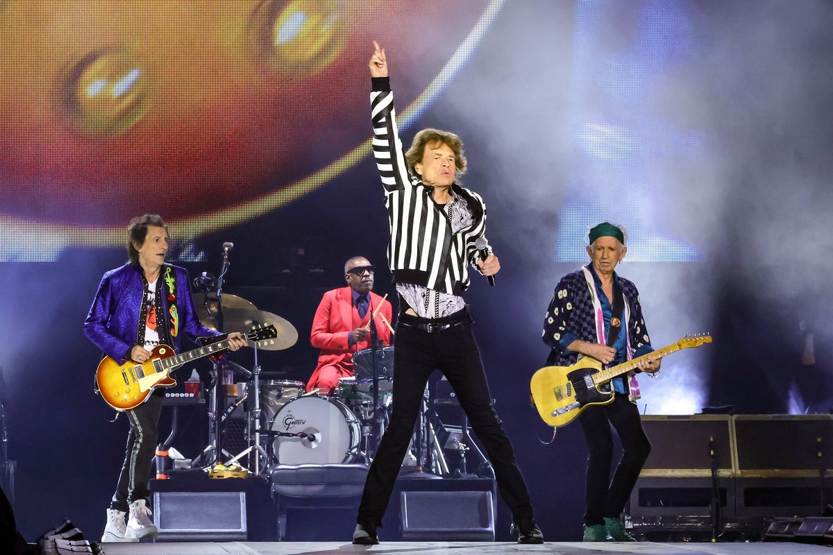 Mick Jagger raises one hand in the air mid-performance alongside The Rolling Stones. He is wearing a black and white striped bomber jacket.