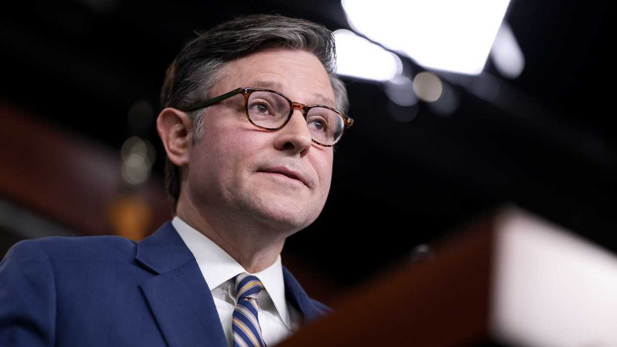 Mike Johnson, a white man with short black hair and glasses, speaking behind a wooden lectern.