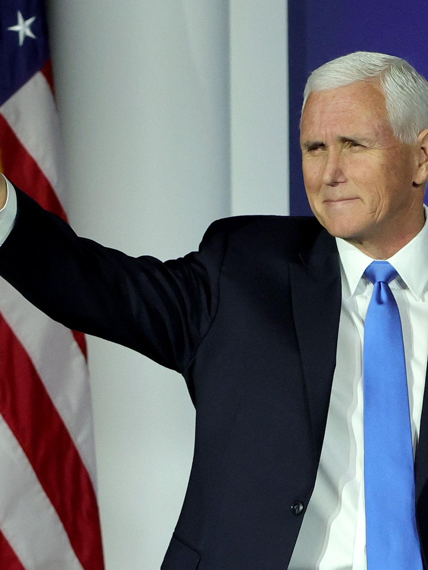 Mike Pence, a white man with short white hair and a black suit, smiles and waves on a stage.