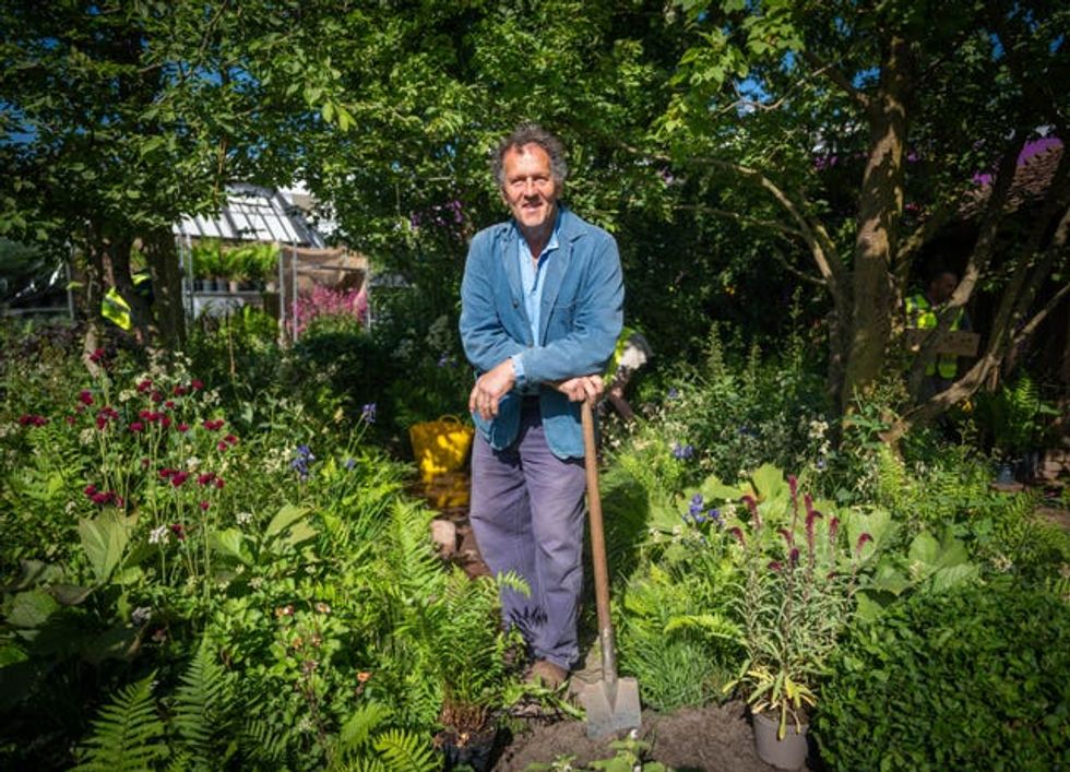 Monty Don, in blue jacket and shirt and grey blue trousers leans on a spade, surrounded by plants
