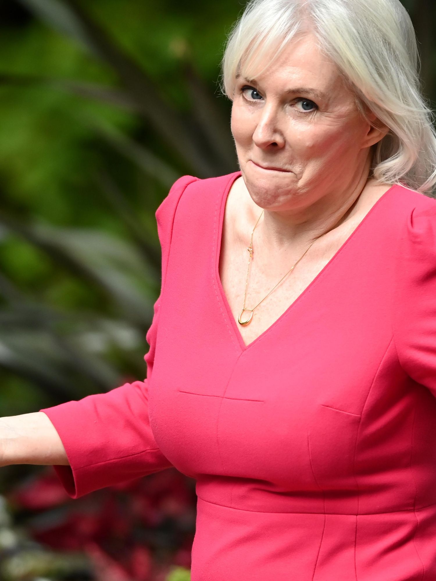 Nadine Dorries, a white woman with long blonde hair and a pink dress, shrugs with her arms out in front of her while walking down Downing Street. She has a red iPhone in her right hand.
