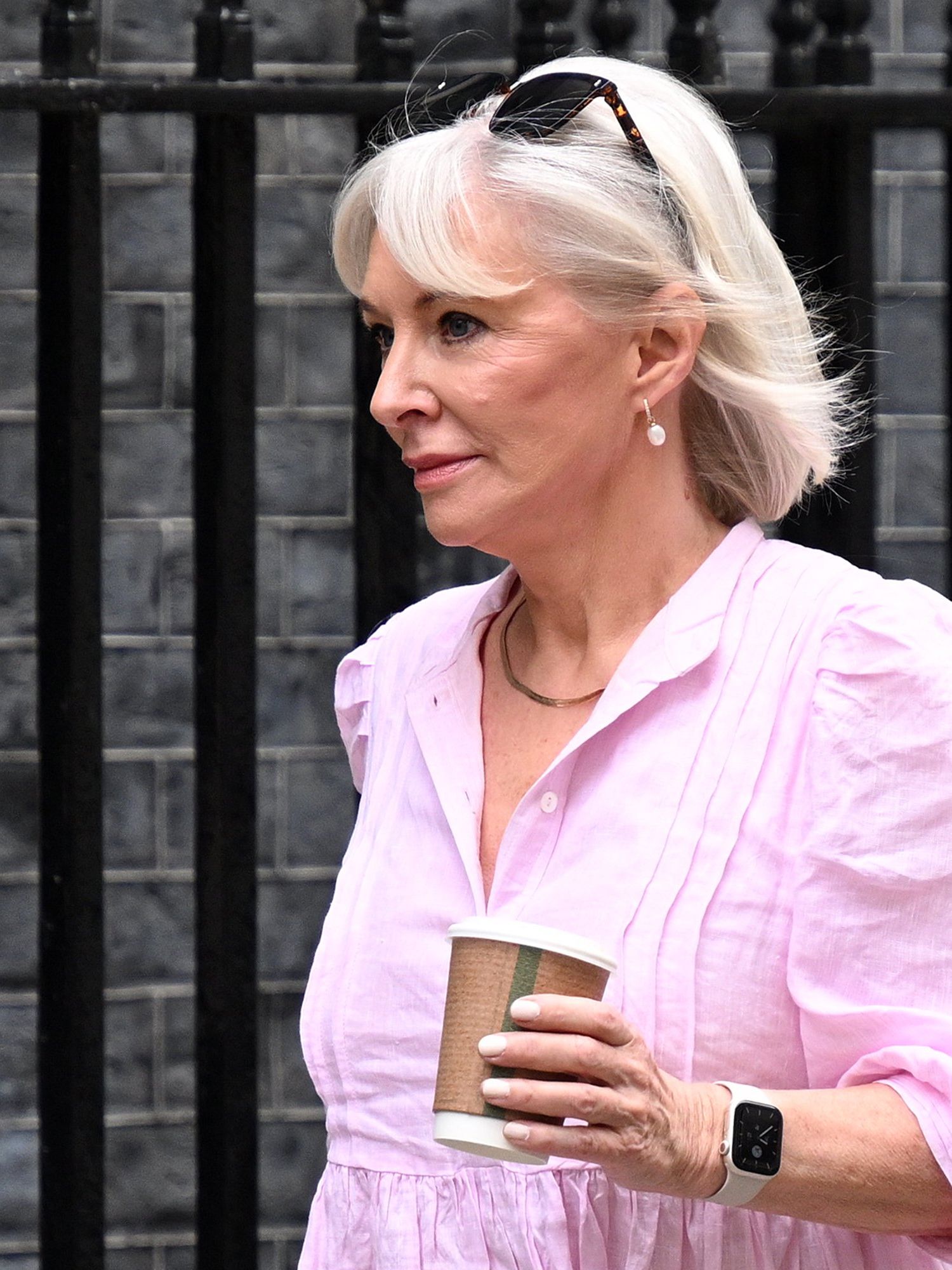 Nadine Dorries, a white woman with white hair and a pink shirt, holds a coffee cup in her hand as she walks down Downing Street.