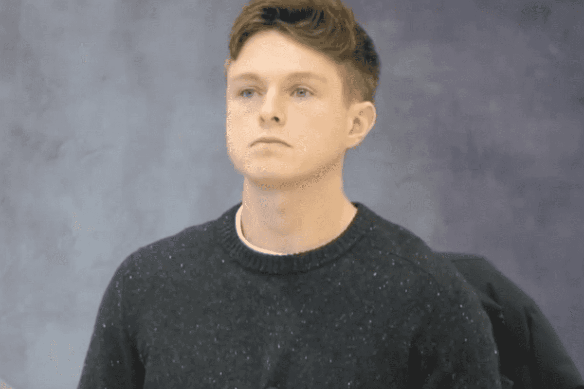 Nathan Cavanaugh, a white man with short brown hair and a black jumper, sitting in a chair against a grey background with a neutral expression on his face.