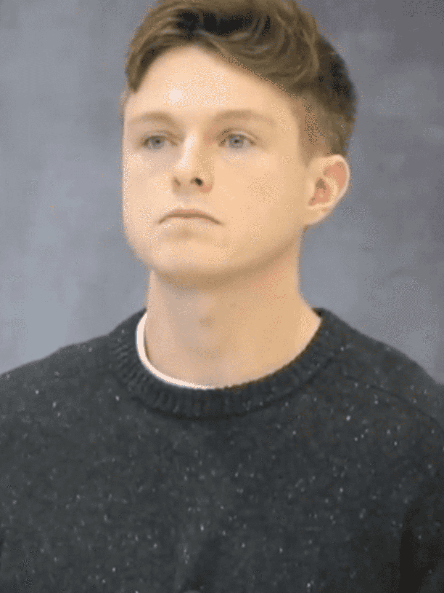 Nathan Cavanaugh, a white man with short brown hair and a black jumper, sitting in a chair against a grey background with a neutral expression on his face.