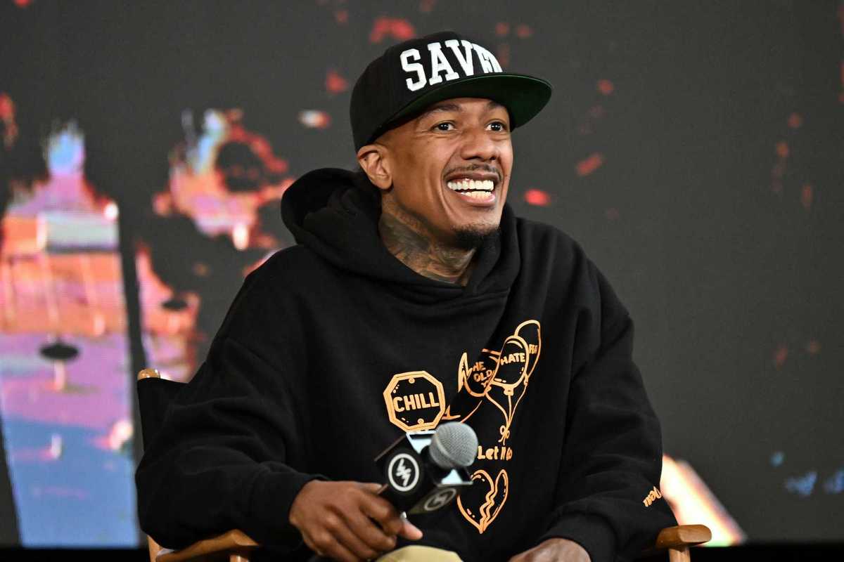 Nick Cannon, a Black man with a black hoodie and black cap which reads 'Saved', sits in a chair and holds a microphone.