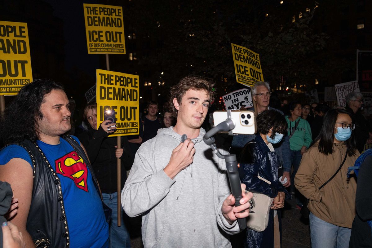 Nick Shirley, a white man with short brown hair and a grey hoodie, holds an iPhone on a gimbal. He's at a protest, with yellow placards reading 'The people demand ICE out' seen behind him.