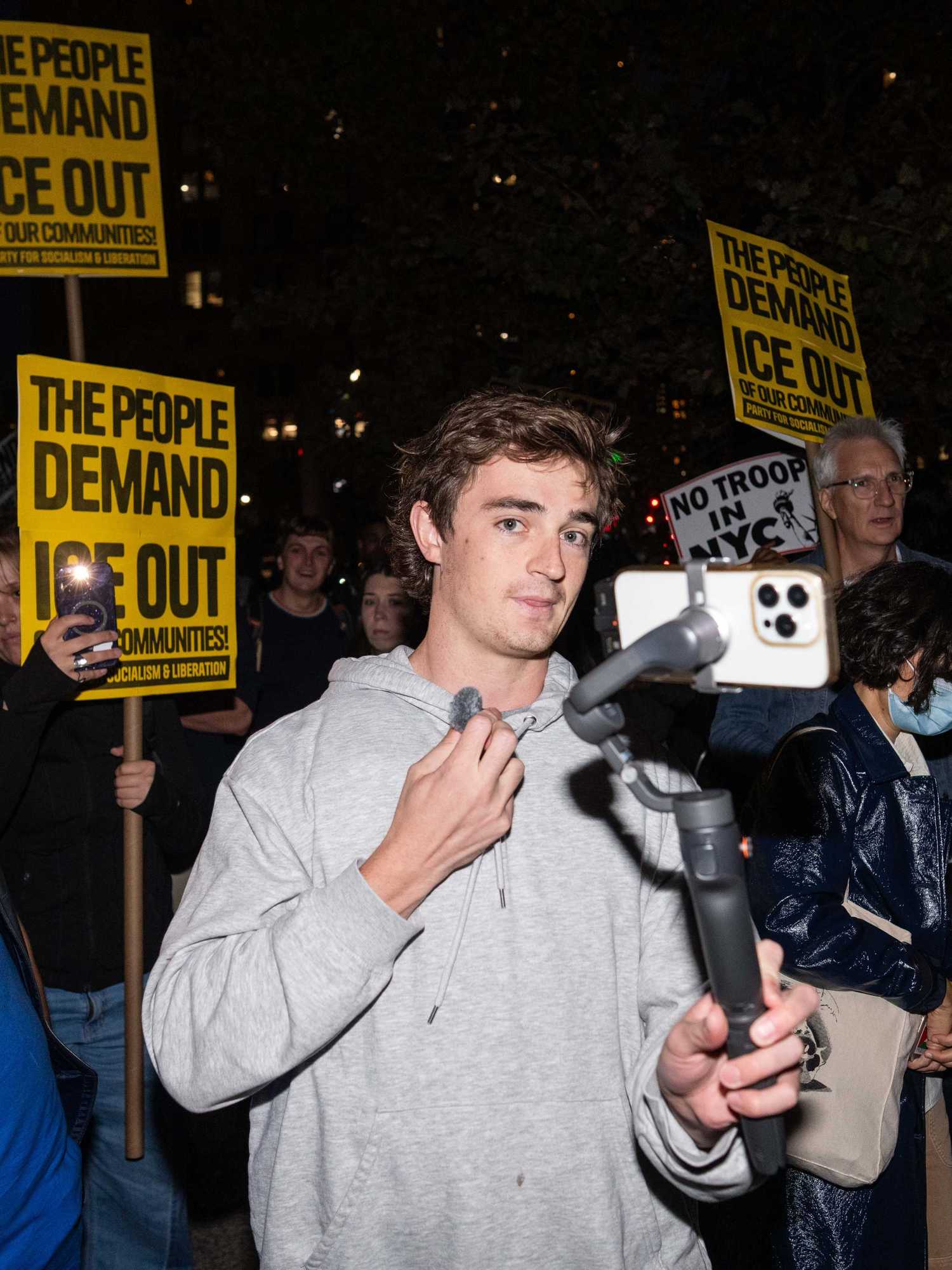 Nick Shirley, a white man with short brown hair and a grey hoodie, holds an iPhone on a gimbal. He's at a protest, with yellow placards reading 'The people demand ICE out' seen behind him.