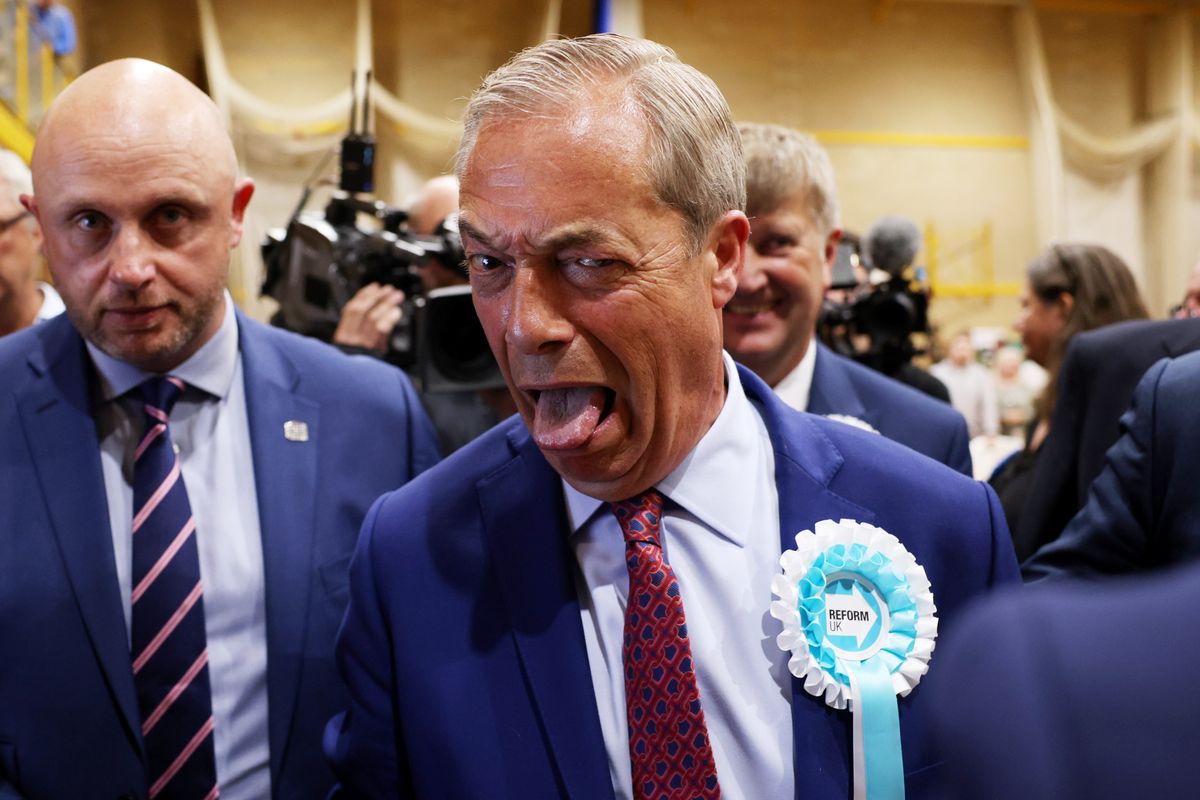 Nigel Farage pulls a face and sticks his tongue out at the election declaration in Clacton. He wears a light blue and white rosette on a blue suit.