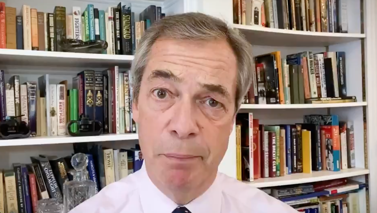 Nigel Farage sits in front of several bookshelves, wearing a lilac collared shirt and spotted blue tie.