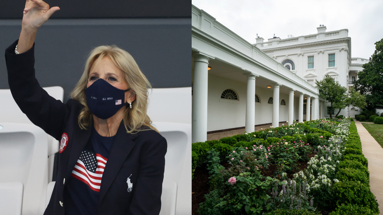 On the left, Jill Biden, a blonde white woman in a black jacket, blue top with the USA flag and a navy face mask, waves. On the right, the White House Rose Garden with shrubbery.