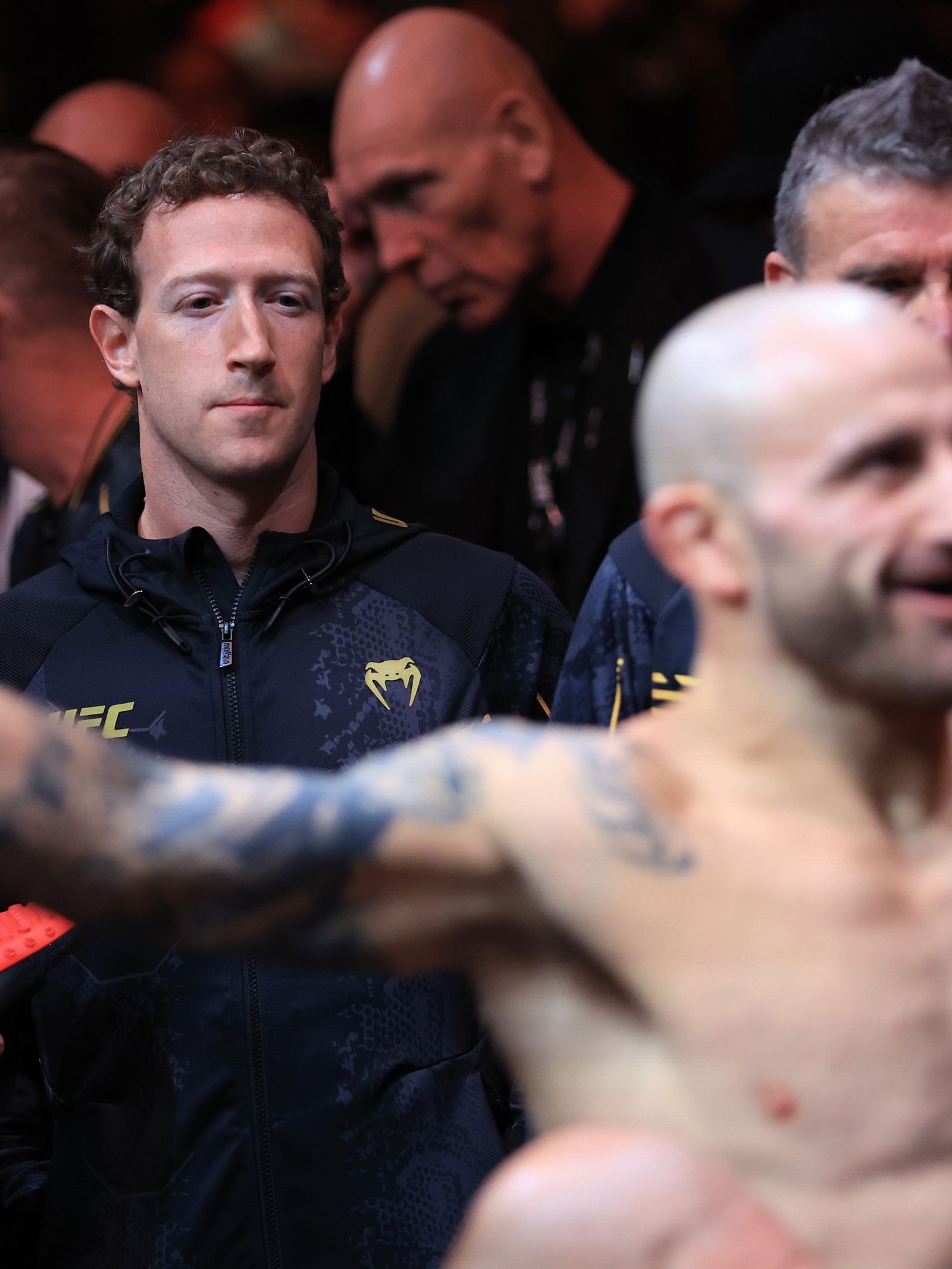 Out of focus in the foreground is Alexander Volkanovski, a bald white man with his arms streched wide. In focus behind him is Mark Zuckerberg, a white man with curly brown hair, closely focussed on Volkanovski and wearing a dark blue zip up jacket.
