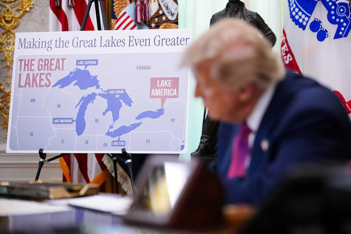 Out of focus in the foreground is Donald Trump signing an executive order at the Resolute Desk. Behind him in focus is a placard showing Lake Ontario renamed as 'Lake America' with the text 'Making the Great Lakes Even Greater'.