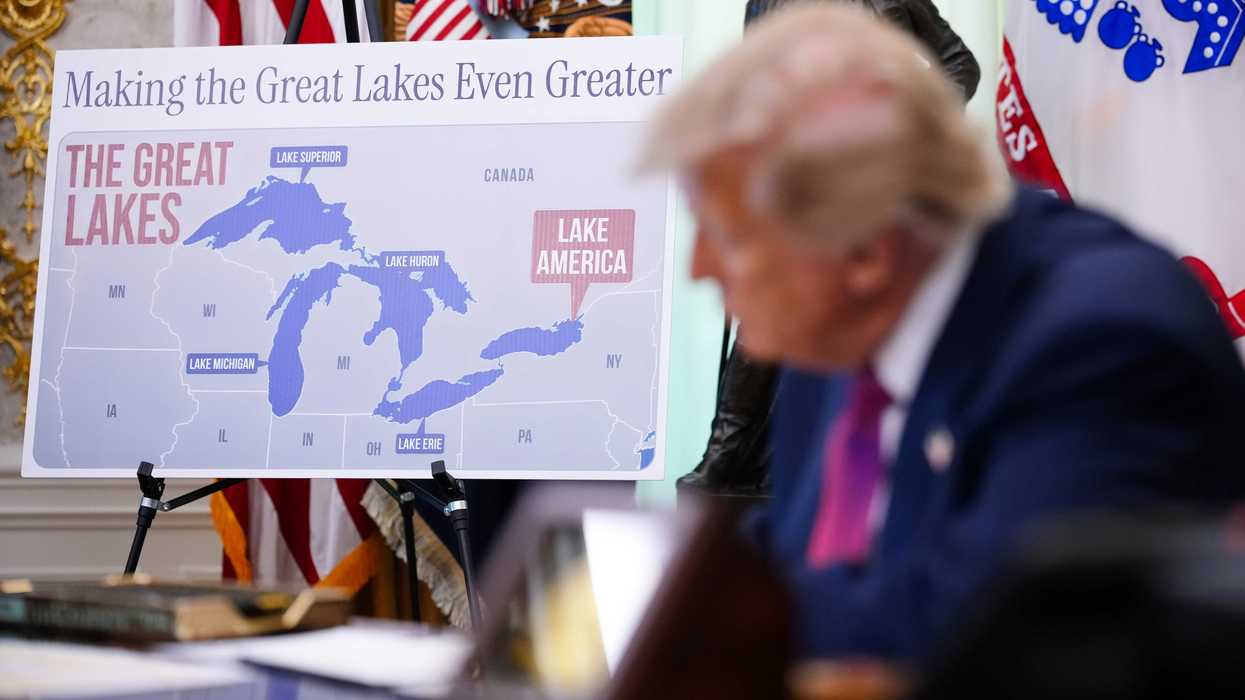 Out of focus in the foreground is Donald Trump signing an executive order at the Resolute Desk. Behind him in focus is a placard showing Lake Ontario renamed as 'Lake America' with the text 'Making the Great Lakes Even Greater'.