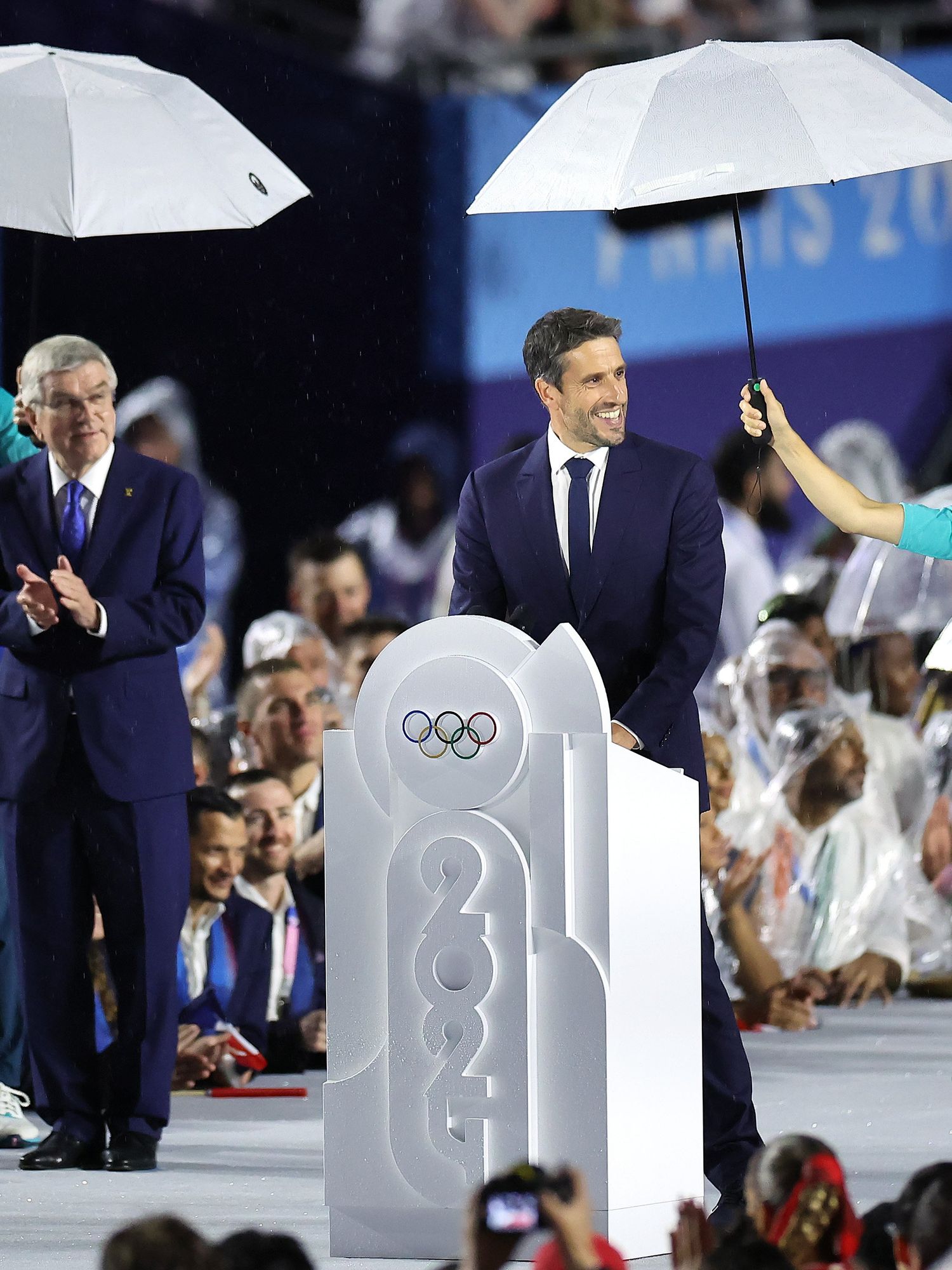 Paris 2024 Organising Committee president Tony Estanguet delivers a speech at the Olympics opening ceremony, with a white woman holding an umbrella next to him. To his right, International Olympic Committee president Thomas Bach watches on, with a white man holding an umbrella for him.