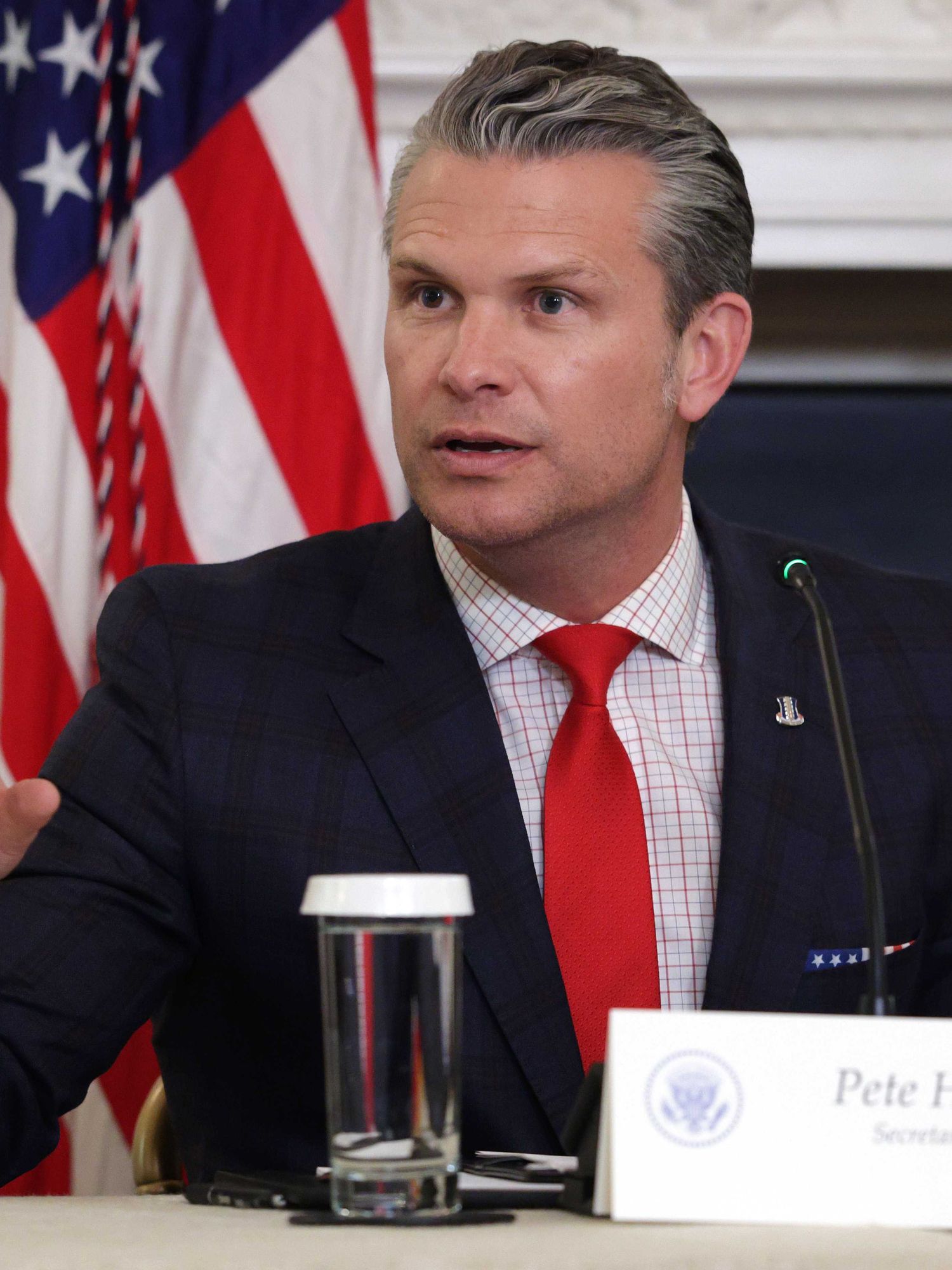 Pete Hegseth, a white man with short grey hair, a black suit and a red tie, talking from beind a desk.