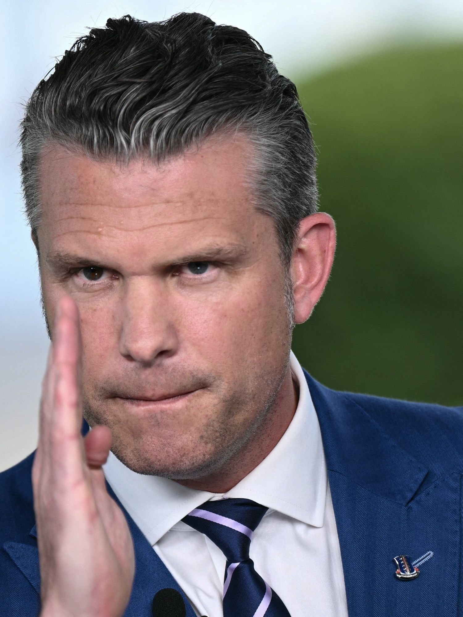Pete Hegseth, a white man with short grey hair and a blue suit, speaking to the media with a neutral facial expression and his right hand in front of him, vertical, with his fingers pointing up.