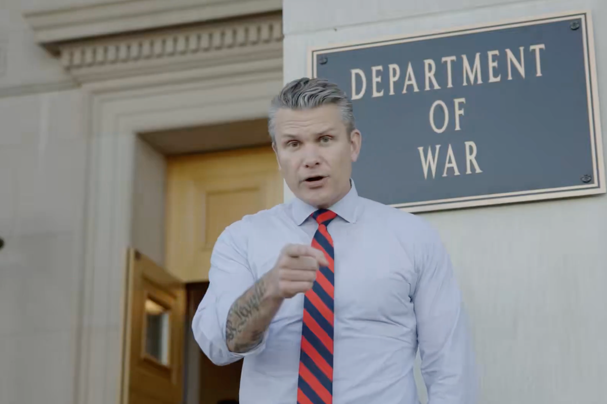 Pete Hegseth, a white man with short grey hair in a white collared shirt and red and blue striped tie, points at the camera as he stands in front of a sign which reads: 'Department of War'.