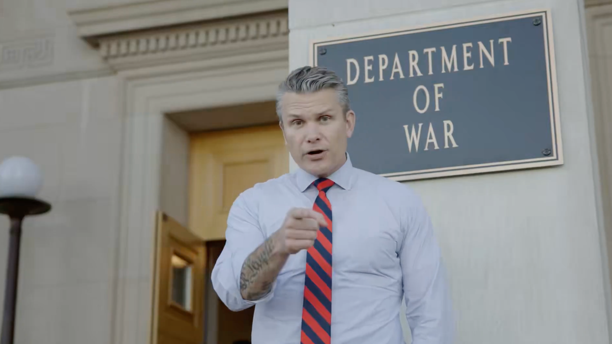 Pete Hegseth, a white man with short grey hair in a white collared shirt and red and blue striped tie, points at the camera as he stands in front of a sign which reads: 'Department of War'.