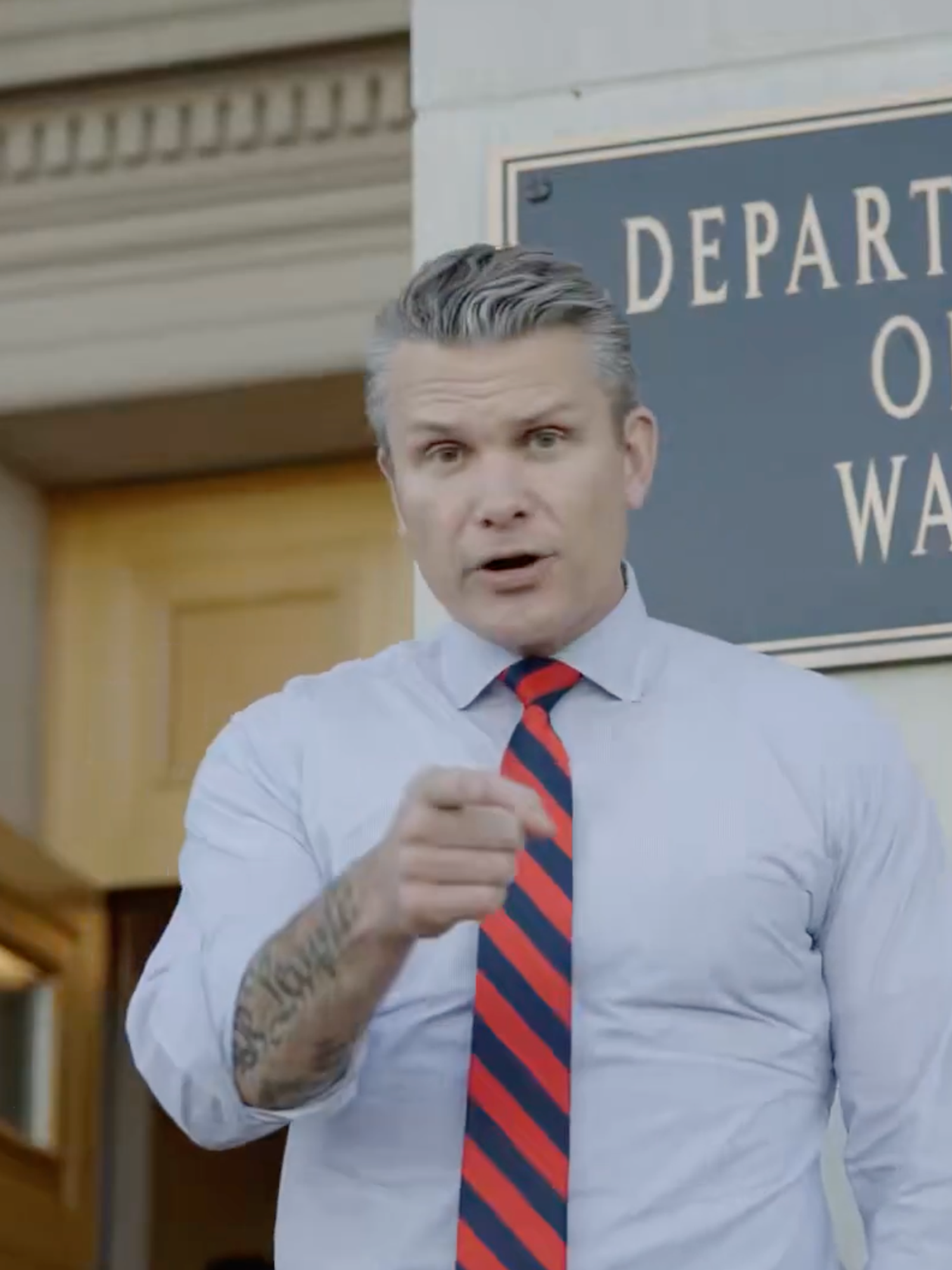 Pete Hegseth, a white man with short grey hair in a white collared shirt and red and blue striped tie, points at the camera as he stands in front of a sign which reads: 'Department of War'.