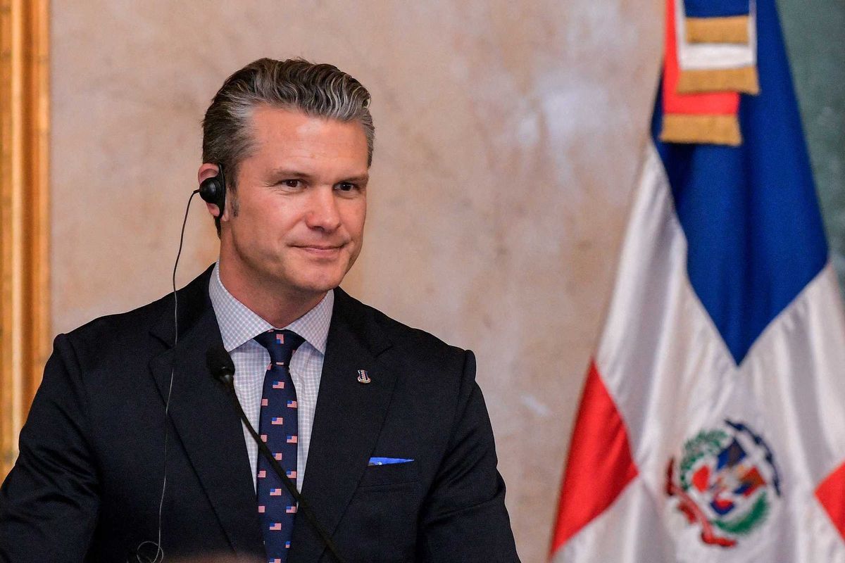 Pete Hegseth in a black suit, wearing a wired translation earpiece in his right ear.