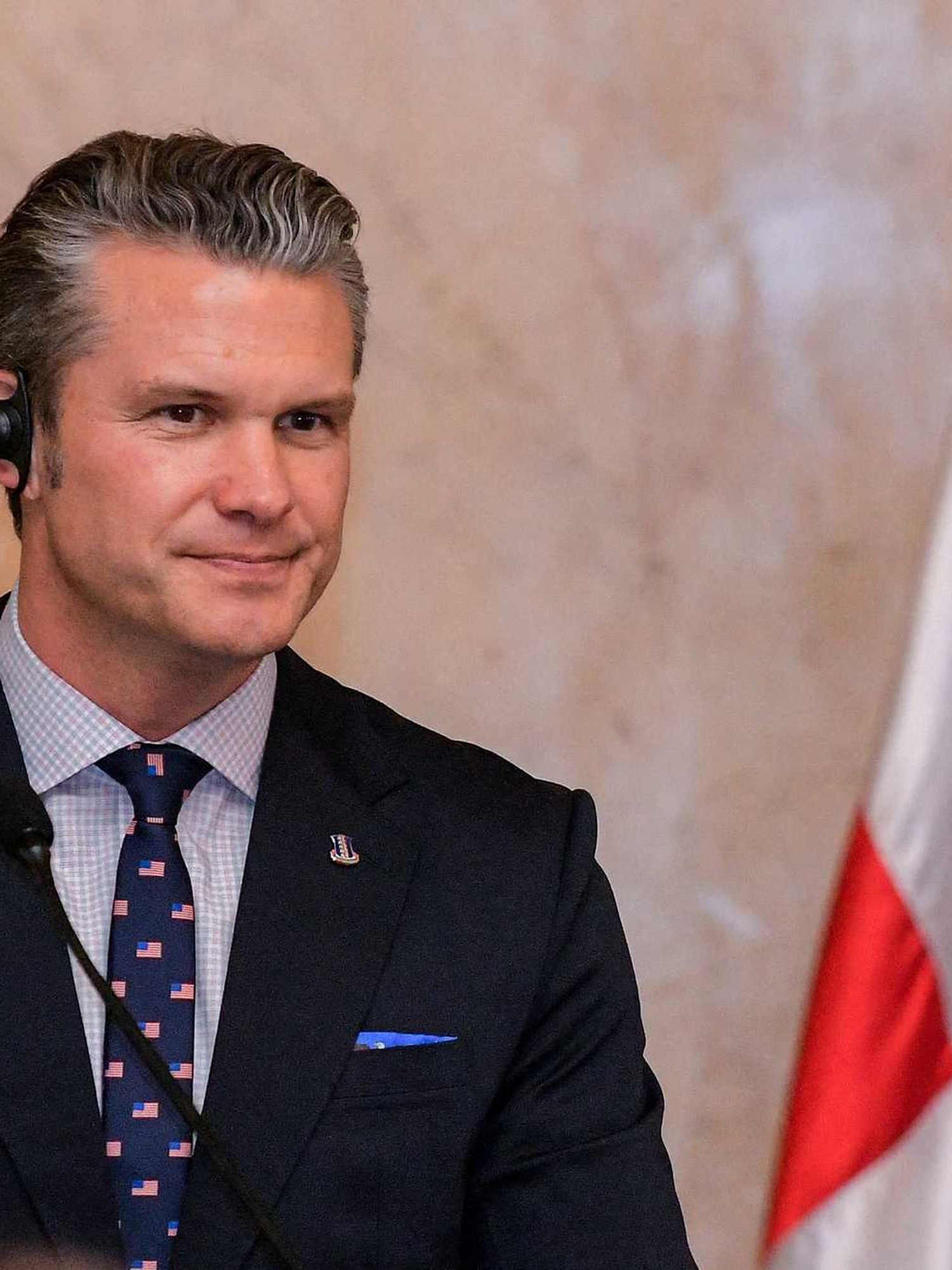 Pete Hegseth in a black suit, wearing a wired translation earpiece in his right ear.