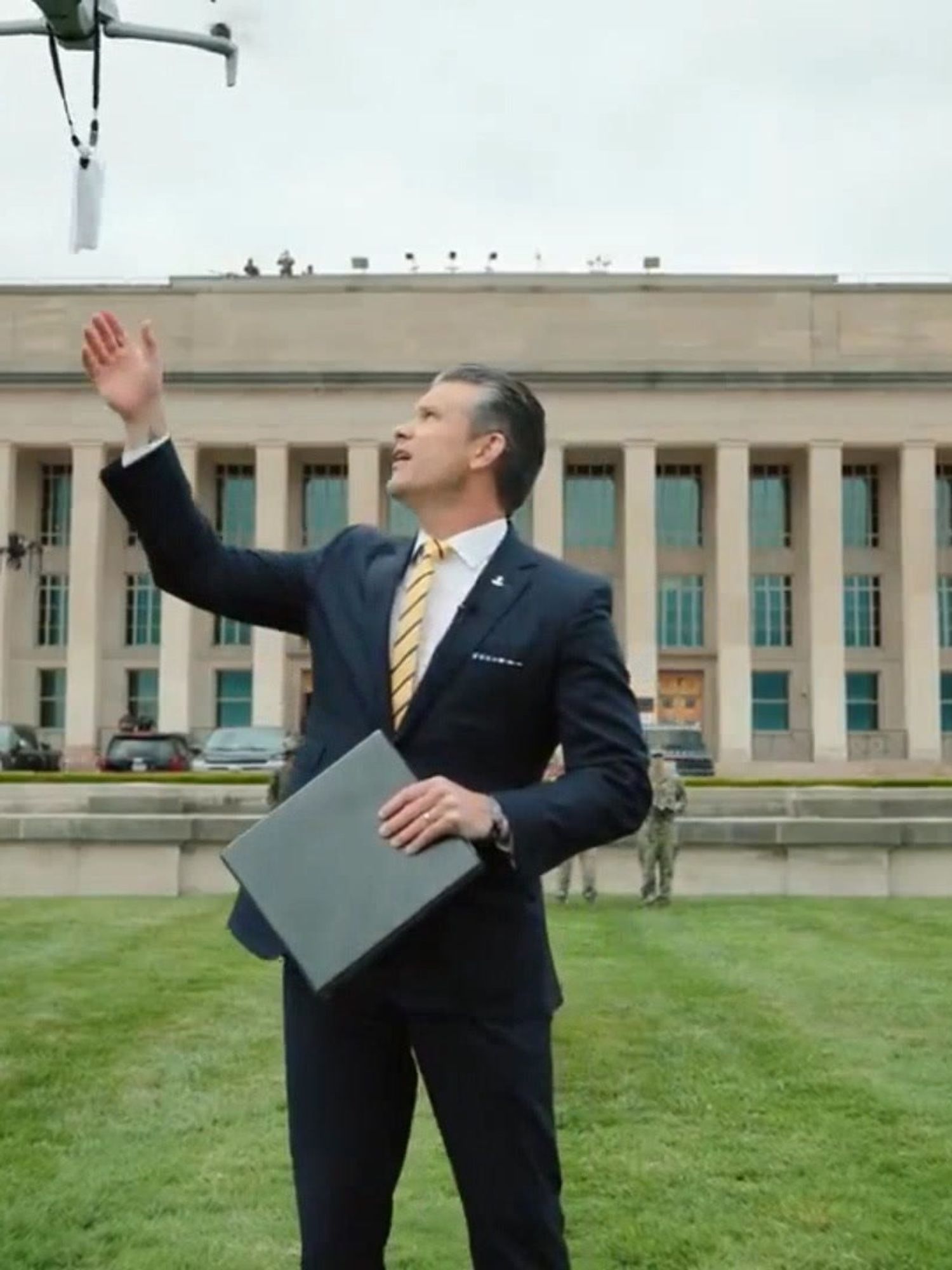 Pete Hegseth outside the Pentagon, holding a file in one hand and reaching for a memo carried by a drone above him in the other.