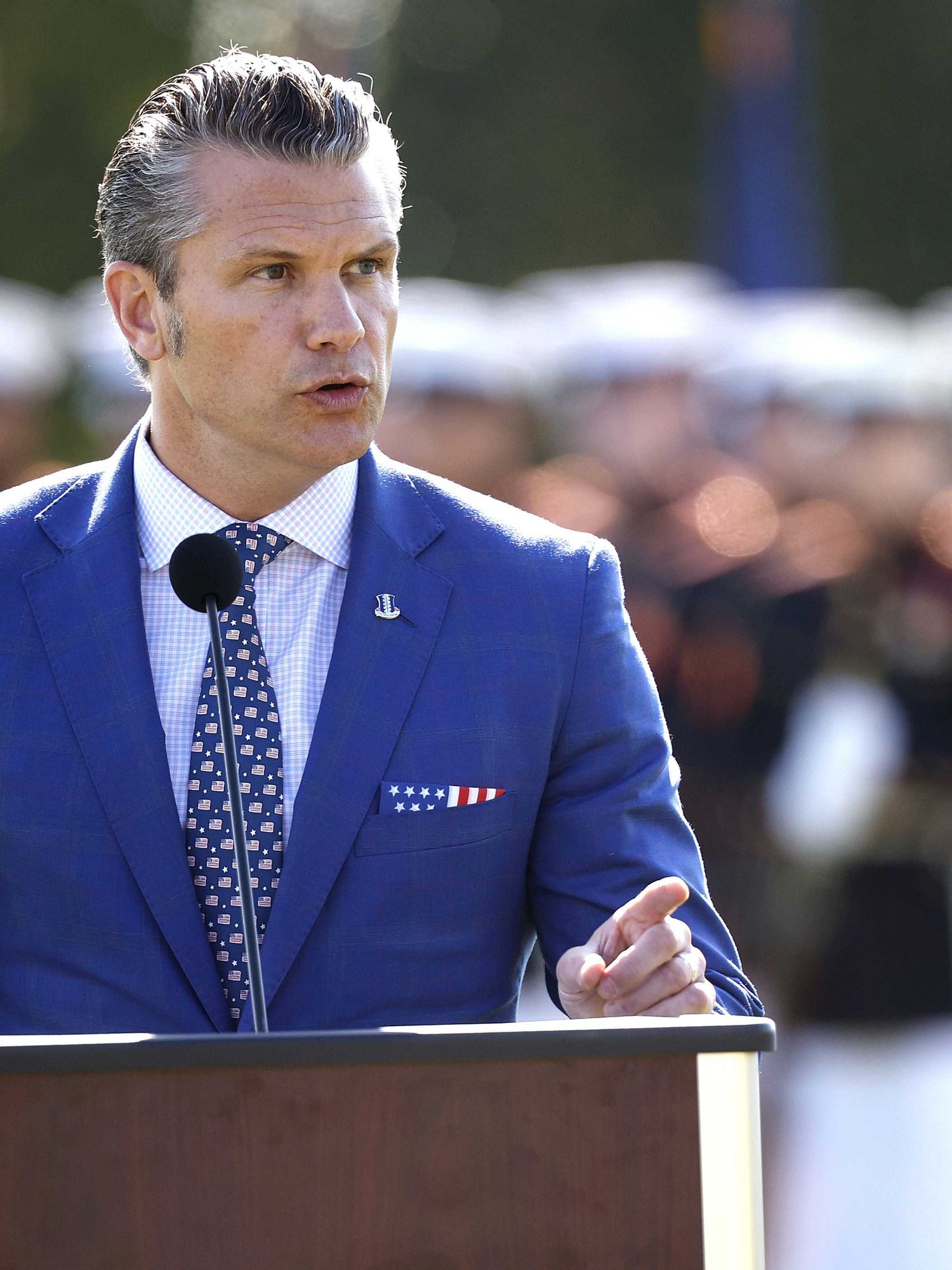 Pete Hegseth speaks behind a wooden lectern, with military seen, out of focus, in the background.