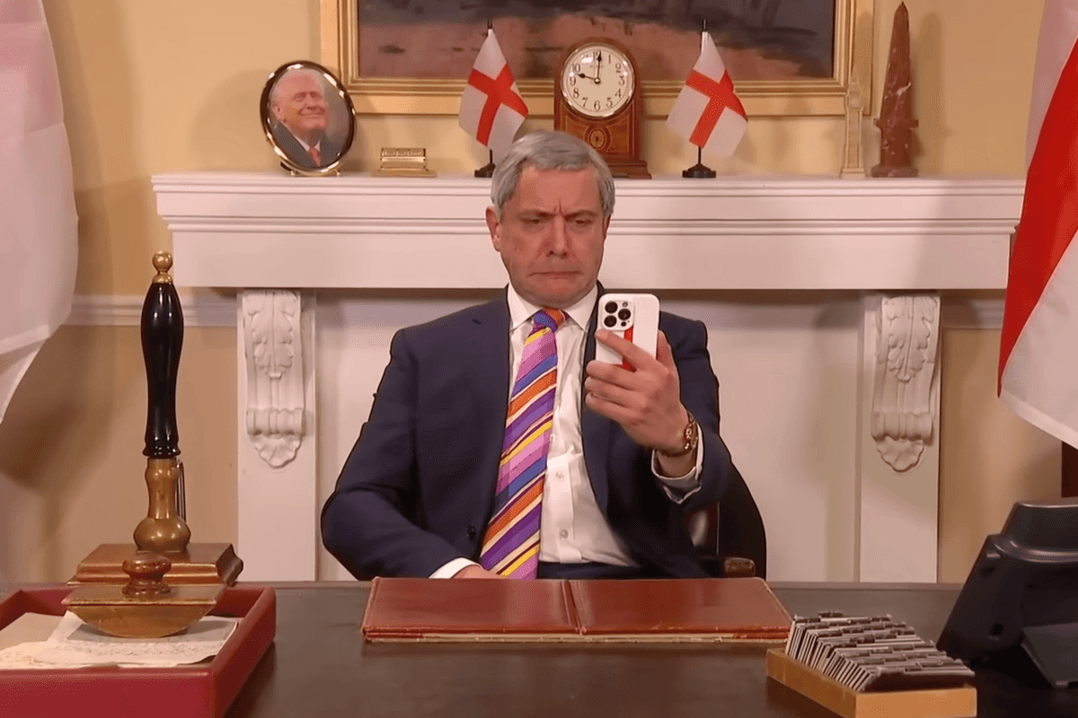 Peter Serafinowicz as Nigel Farage on Saturday Night Live UK, wearing a striped tie and holding up a phone in his left hand.