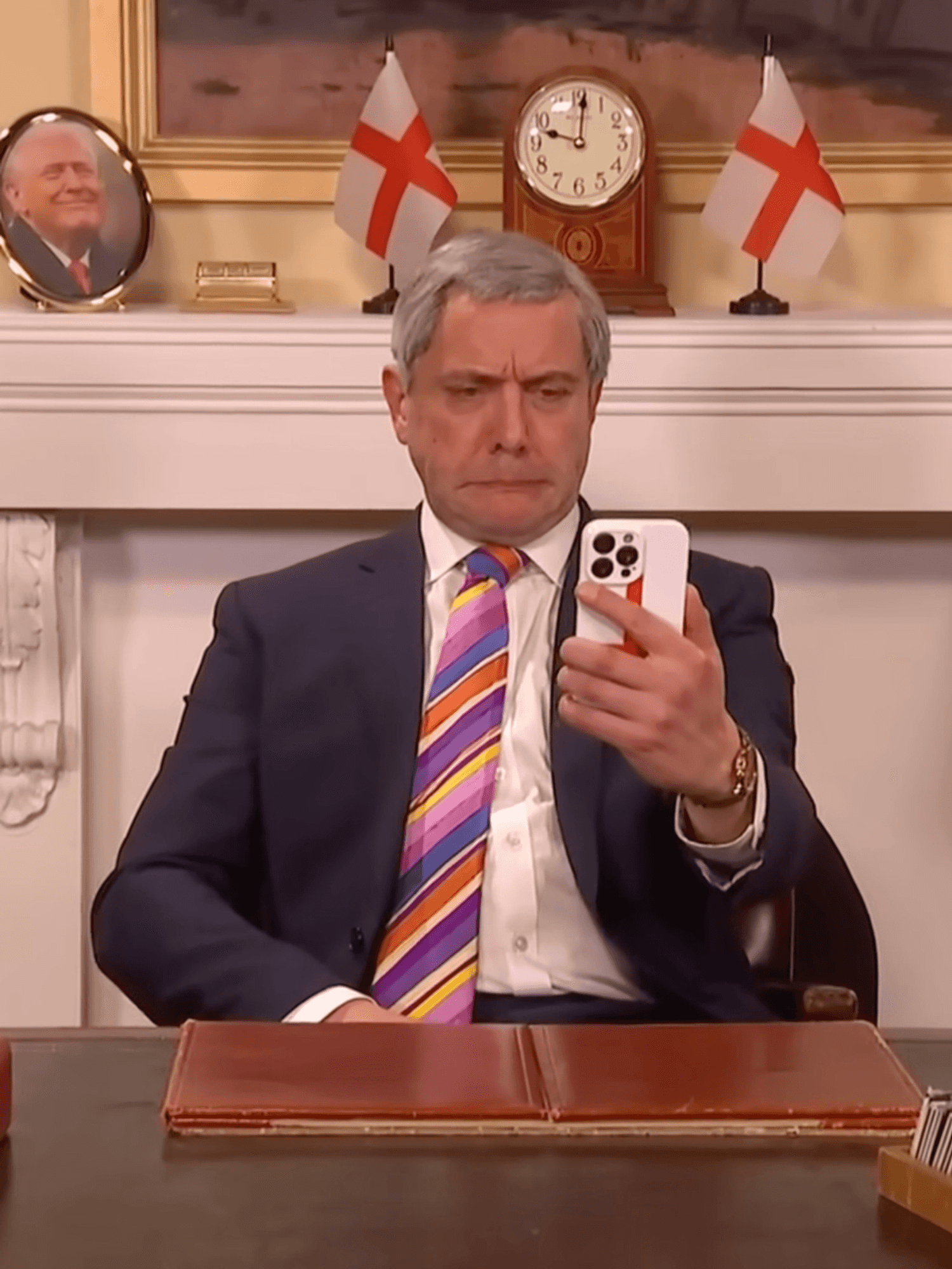Peter Serafinowicz as Nigel Farage on Saturday Night Live UK, wearing a striped tie and holding up a phone in his left hand.