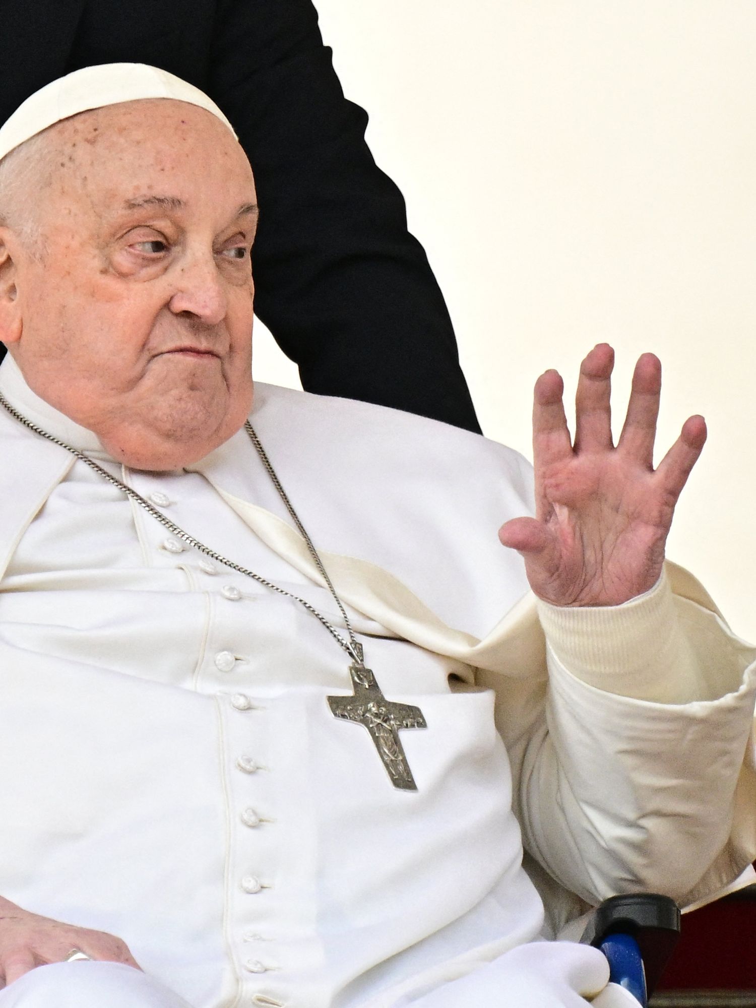 Pope Francis in a wheelchair, waving with his left hand.