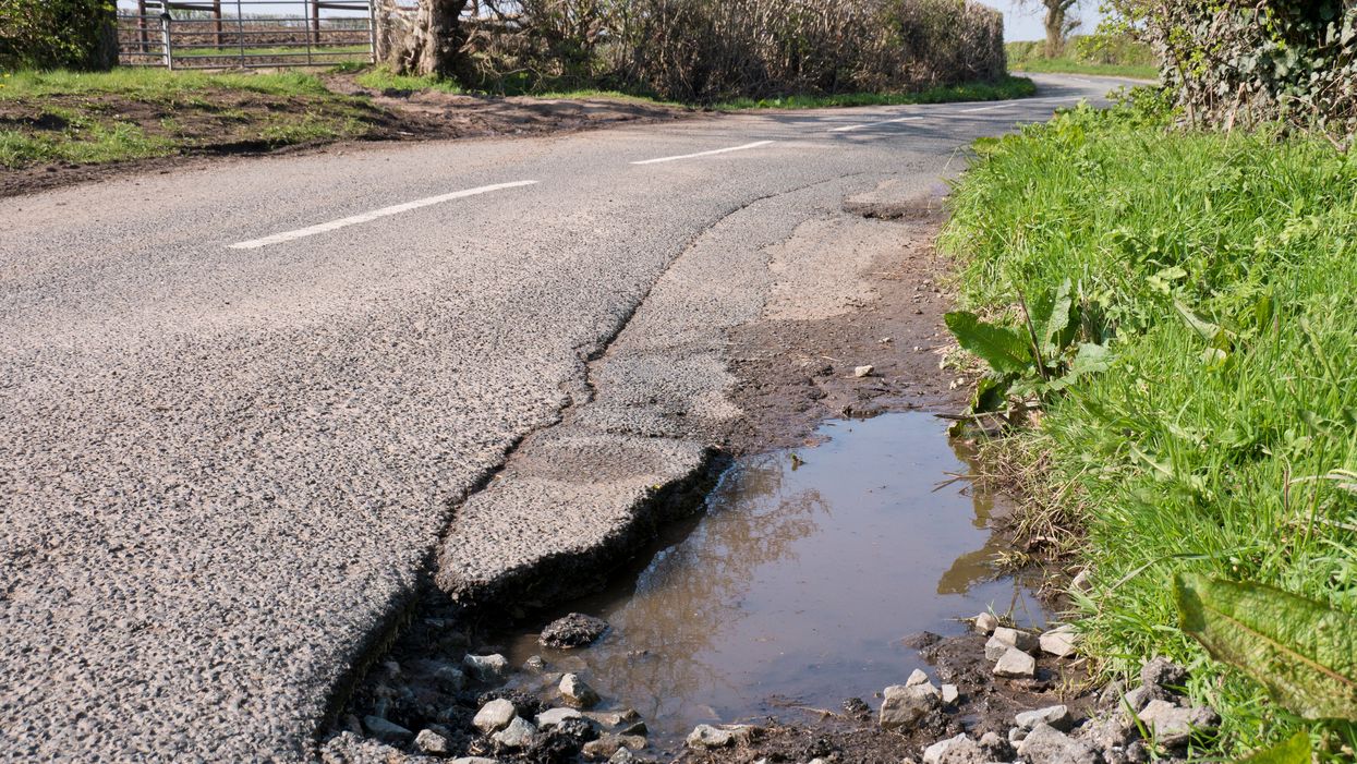 Pothole on the side of a country road, with bushes, green grass and trees on either side of it.