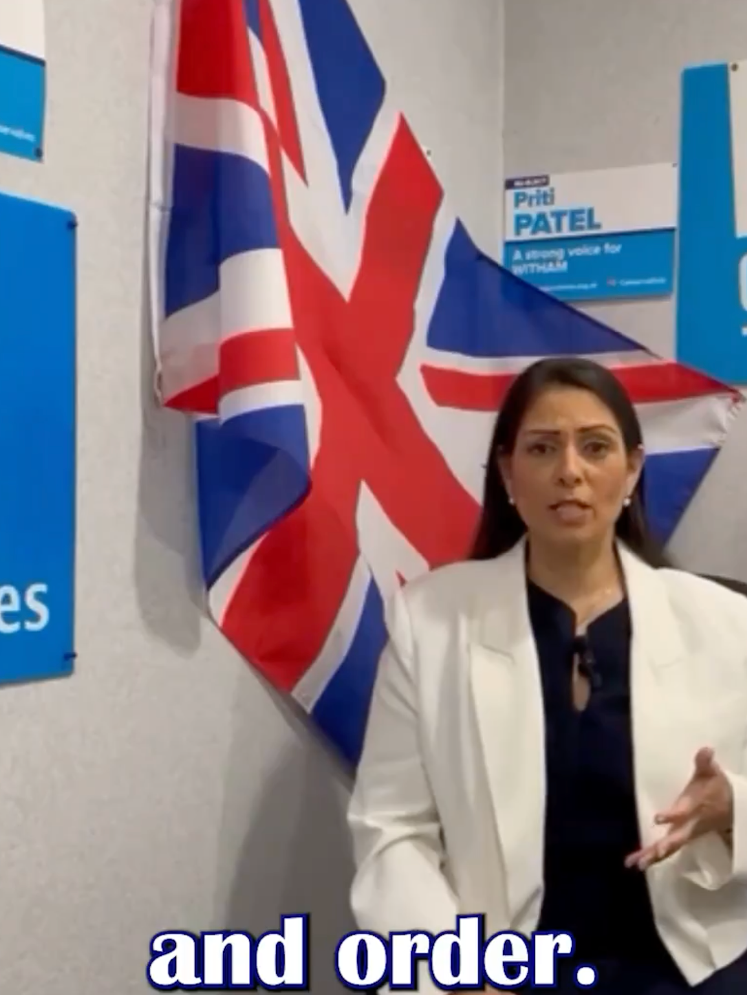 Priti Patel stands in front of a wall adorned with a Union Jack, multiple blue Conservative posters and a framed picture of Boris Johnson.