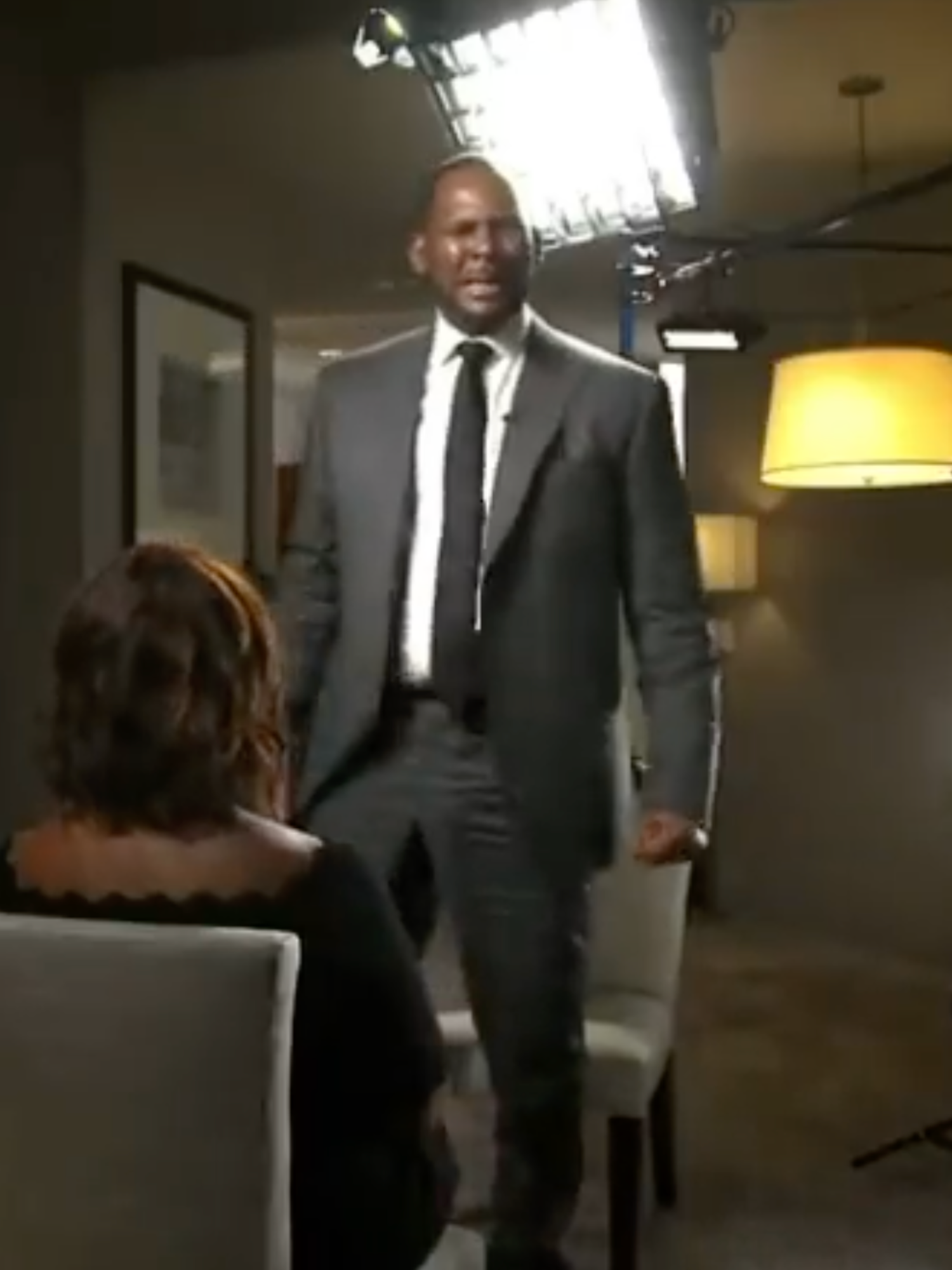 R Kelly, a Black man in a grey suit, stands up amid a TV interview in a hotel room, while a Black woman sits down opposite him. He is emotional.