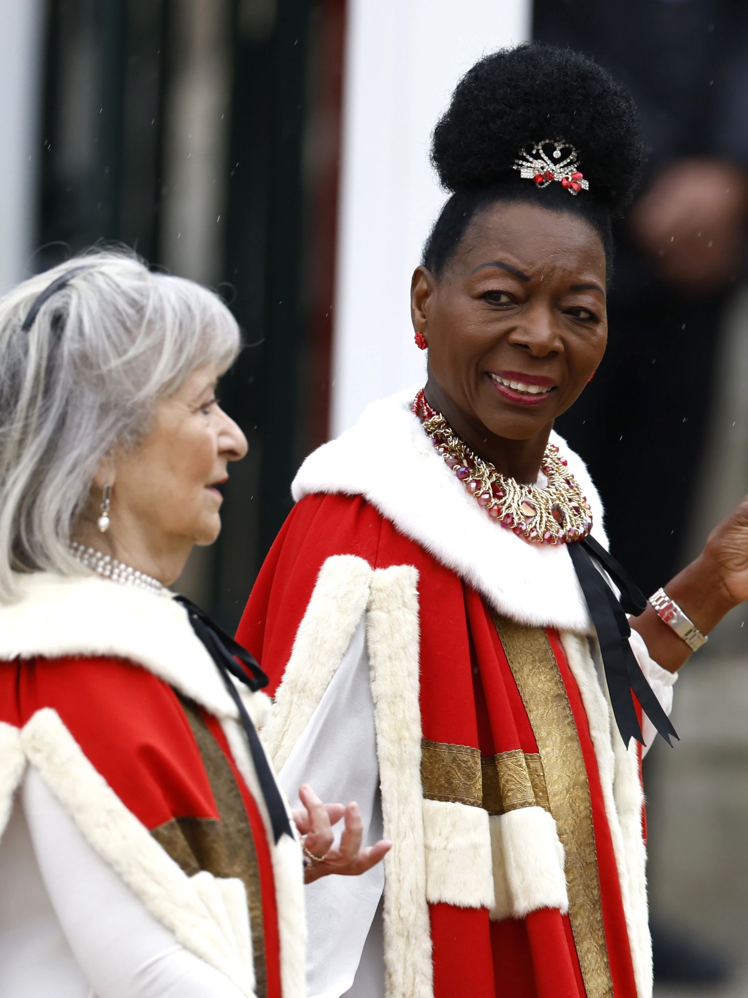 Right, Baroness Floella Benjamin in a red and white cloak walks outside Westminster Abbey. She's a Black woman with her short black hair tied upright.