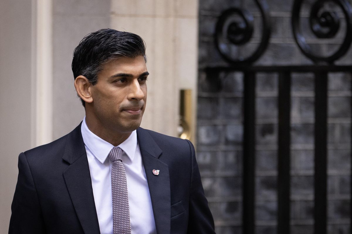 Rishi Sunak, a brown man with short black hair and a black suit, walks out of 10 Downing Street.